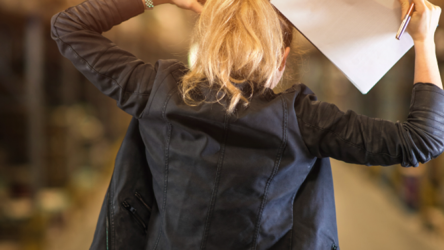 A woman with blonde hair, seen from behind, stands in a warehouse aisle holding a clipboard and pen, scratching her head as if puzzled or thinking. She wears a black jacket and a gray skirt.