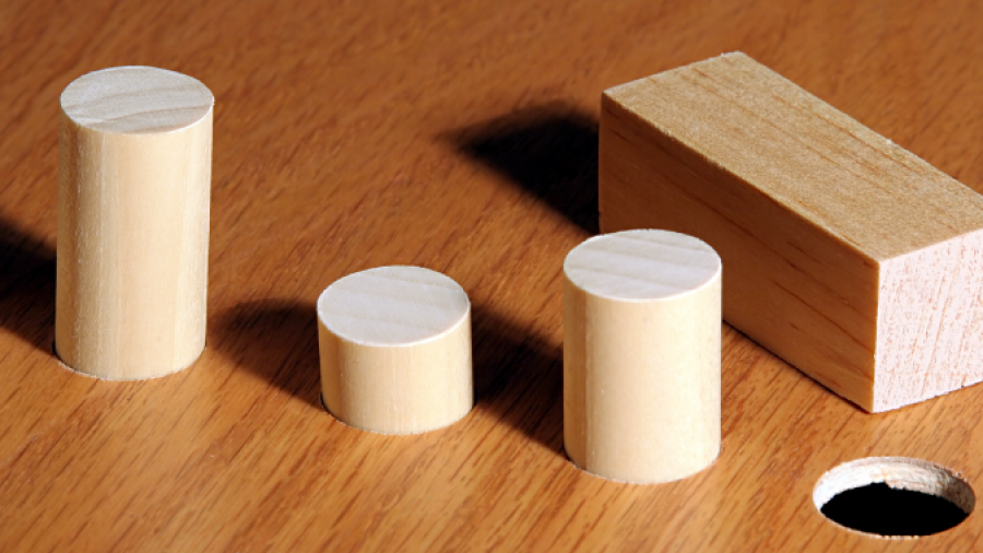 Three wooden cylinders and one rectangular block sit on a wooden surface. There is a round hole in the surface near the cylinders, suggesting a shape-sorting or fitting puzzle.
