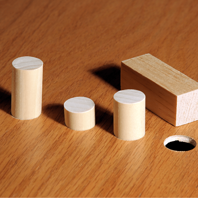 Three wooden cylinders and one rectangular block sit on a wooden surface. There is a round hole in the surface near the cylinders, suggesting a shape-sorting or fitting puzzle.