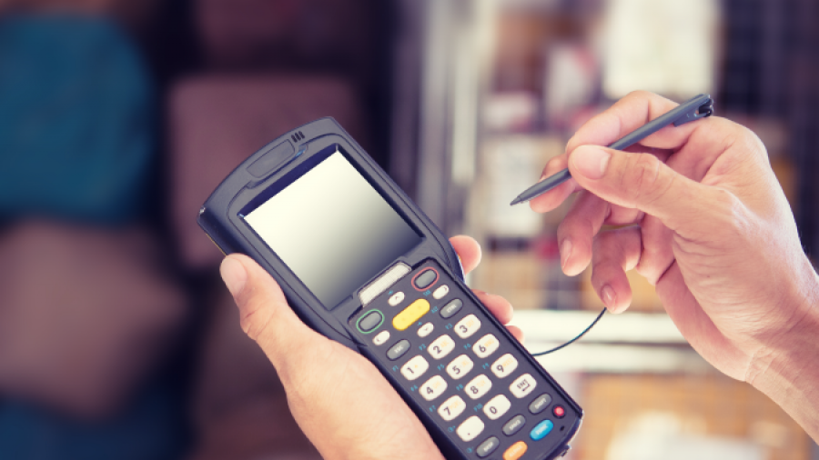 A person uses a stylus on a handheld barcode scanner or inventory device, with shelves and packages blurred in the background.
