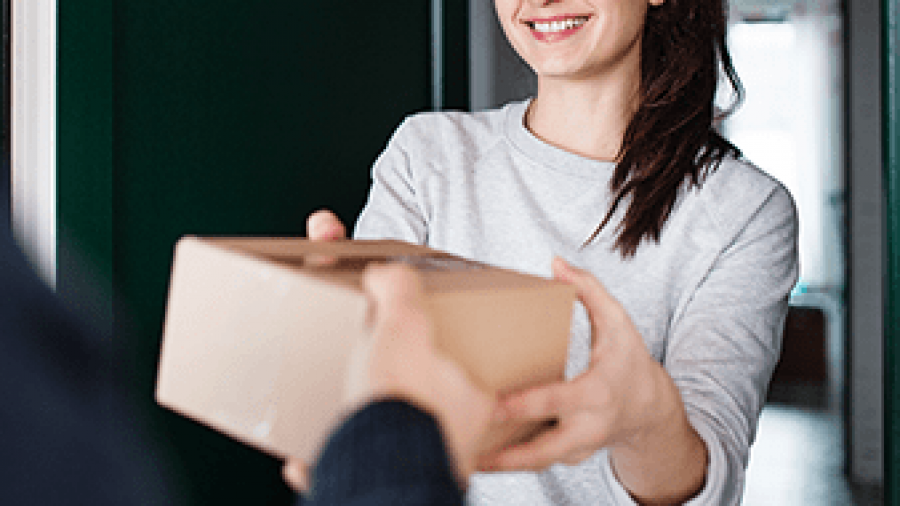 A smiling woman in a light gray sweater hands a cardboard package to another person at a doorway.