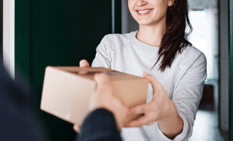 A smiling woman in a light gray sweater hands a cardboard package to another person at a doorway.