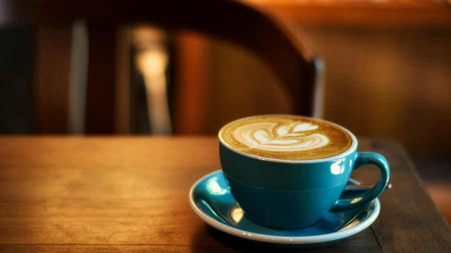 A blue cup of latte with intricate foam art sits on a matching saucer atop a wooden table, evoking the thoughtful ambiance often found in Business Intelligence Consulting spaces, with a blurred chair and bookshelf in the warm, softly lit background.