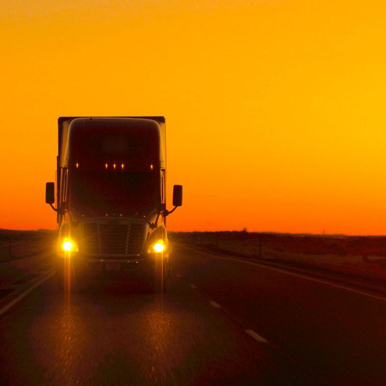 A large semi-truck with headlights on drives down a highway at sunset, silhouetted against an orange and yellow sky.
