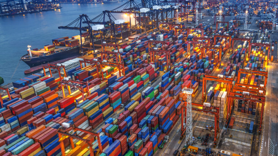 A busy shipping port at dusk with rows of colorful cargo containers, large cranes, and ships docked along the water, illuminated by bright lights. City buildings are visible across the river in the background.