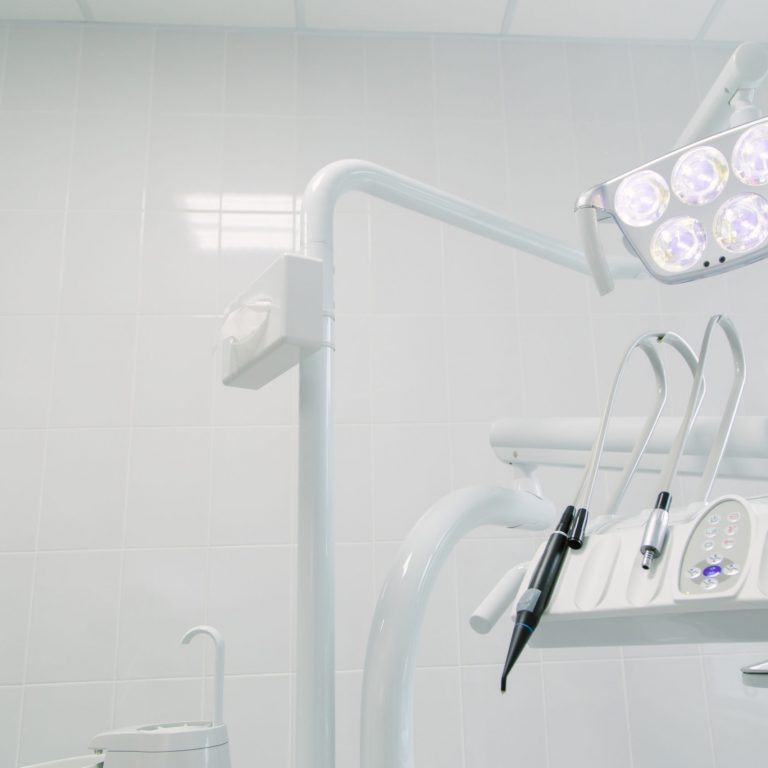 A close-up of modern dental equipment in a brightly lit, clean dental clinic, featuring overhead lights and various dental tools against a white tiled wall.