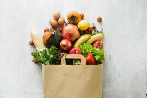 A paper grocery bag overflows with fresh produce, bread, leafy greens, apples, bananas, eggs, pomegranate, squash, tomatoes, strawberries, and other colorful fruits and vegetables against a light background.