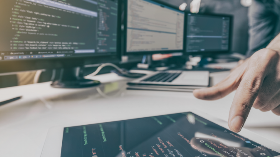 A person uses a tablet displaying code, with multiple computer monitors showing programming scripts on a modern desk in a tech workspace.
