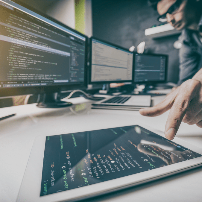 A person uses a tablet displaying code, with multiple computer monitors showing programming scripts on a modern desk in a tech workspace.