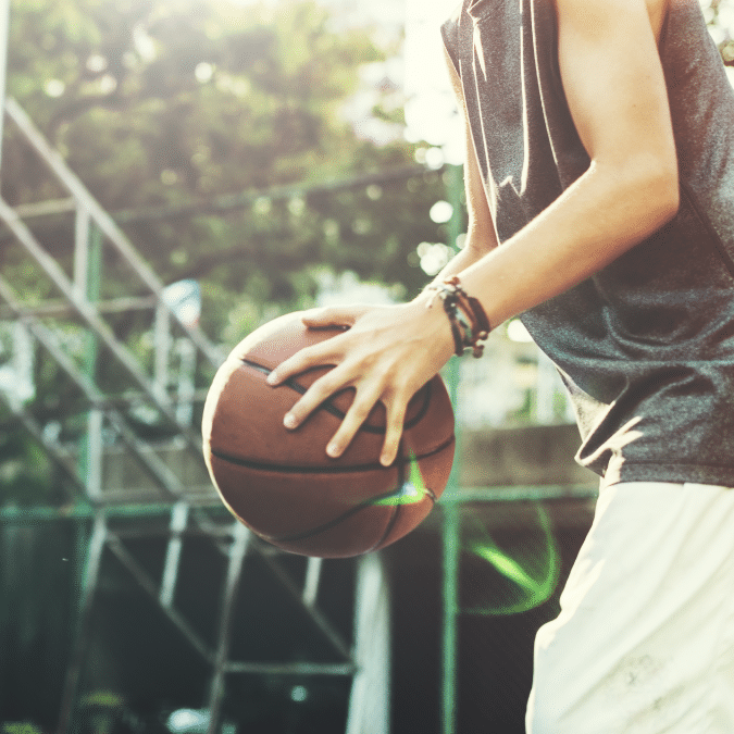 A person wearing a sleeveless shirt and shorts dribbles a basketball outdoors on a sunny day, with a hoop and a metal fence visible in the background.