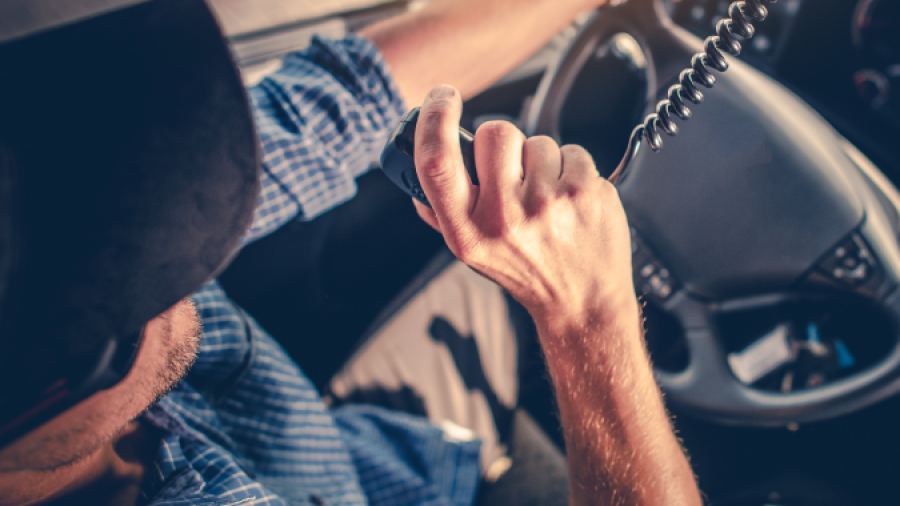 A person wearing a cap and blue plaid shirt drives a vehicle while speaking into a CB radio, with one hand on the steering wheel and sunlight streaming through the window.