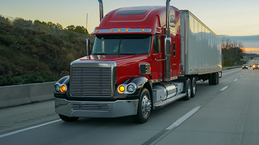 A red semi-truck with a large white trailer drives down a highway at dusk, passing green trees and grassy areas along the roadside under a clear blue sky.