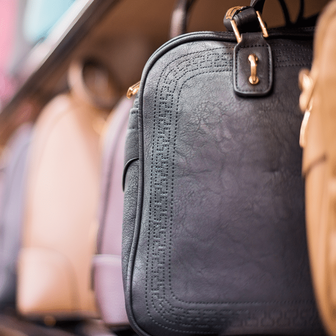 A close-up of a black leather handbag with gold hardware on a shelf, surrounded by other blurred handbags in various neutral colors.