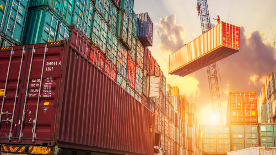 Shipping containers stacked high at a busy port, with a crane lifting a container and a truck parked nearby. The sun sets in the background, casting a warm glow—an ideal scene for showcasing efficient consumer goods consulting in global trade.
