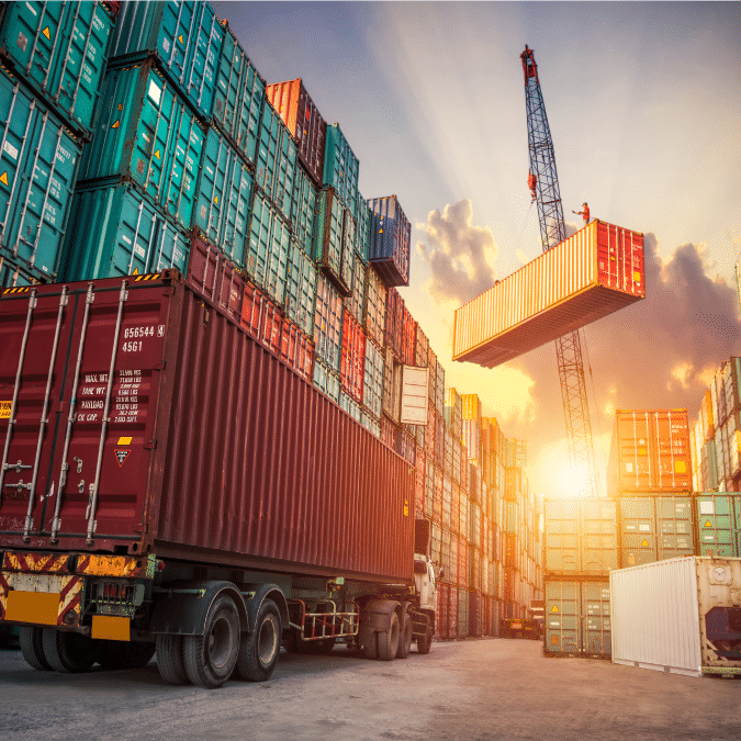 Shipping containers stacked high at a busy port, with a crane lifting a container and a truck parked nearby. The sun sets in the background, casting a warm glow—an ideal scene for showcasing efficient consumer goods consulting in global trade.