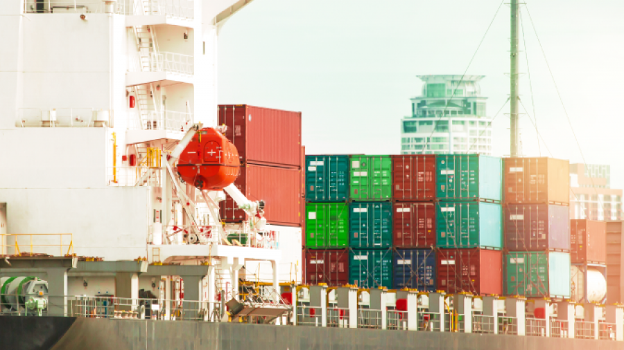 A cargo ship docked at a port, loaded with colorful shipping containers, with city buildings visible in the background.