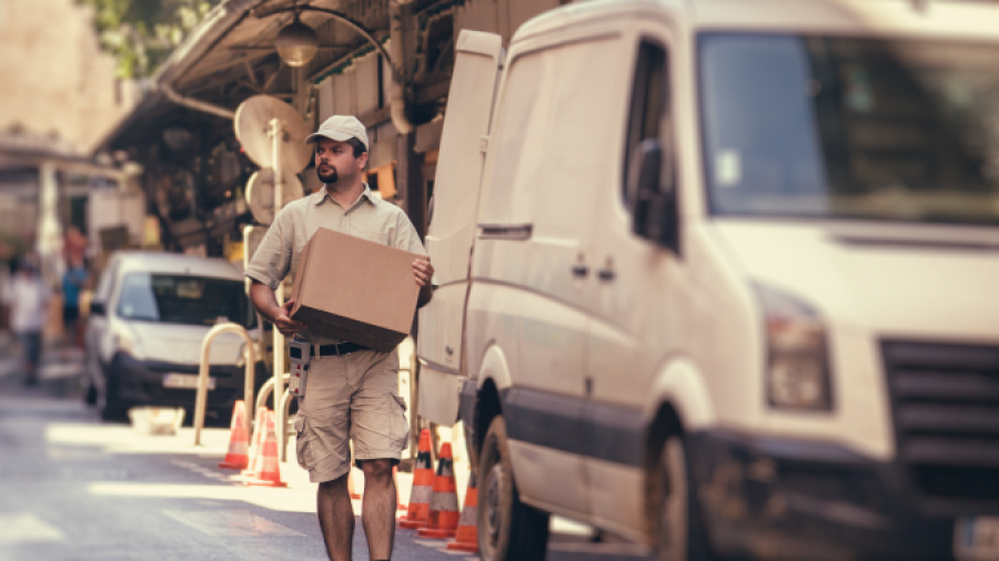 A delivery person in a khaki uniform and cap carries a cardboard box along a city street beside a white van parked by the curb on a sunny day. Traffic cones are set up near the vehicles.