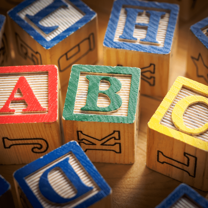Colorful wooden alphabet blocks featuring raised letters A, B, and D in the foreground, with several other blocks displaying different letters in the background, all placed on a wooden surface.