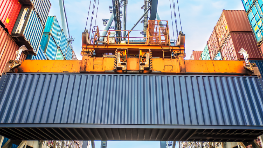 A large crane lifts a blue shipping container between stacks of colorful containers at a busy port, with metal structures and a clear sky in the background.