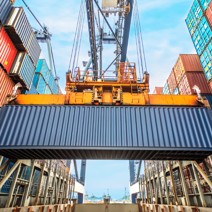 A large crane lifts a blue shipping container between stacks of colorful containers at a busy port, with metal structures and a clear sky in the background.