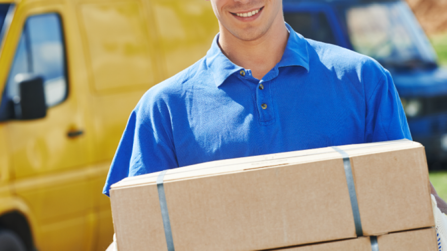 A smiling delivery person in a blue uniform and cap carries two cardboard boxes, standing in front of a yellow and blue delivery van outdoors.