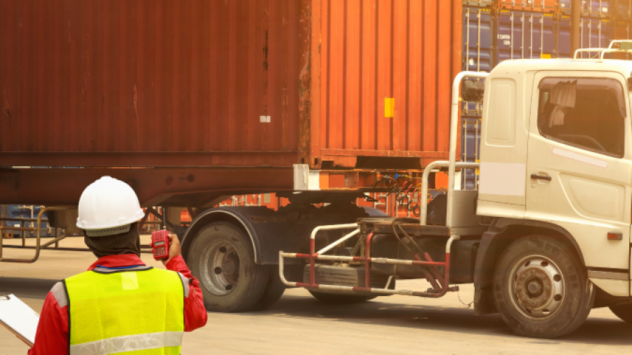 A worker in a safety vest and helmet holds a clipboard and gestures toward a large shipping container being transported by a truck, with stacked containers in the background.