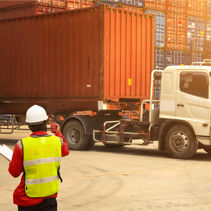 A worker in a safety vest and helmet holds a clipboard and gestures toward a large shipping container being transported by a truck, with stacked containers in the background.