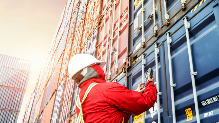 A worker wearing a red safety suit, helmet, and reflective vest inspects or secures stacks of shipping containers outdoors at a shipping yard, with sunlight illuminating the scene.