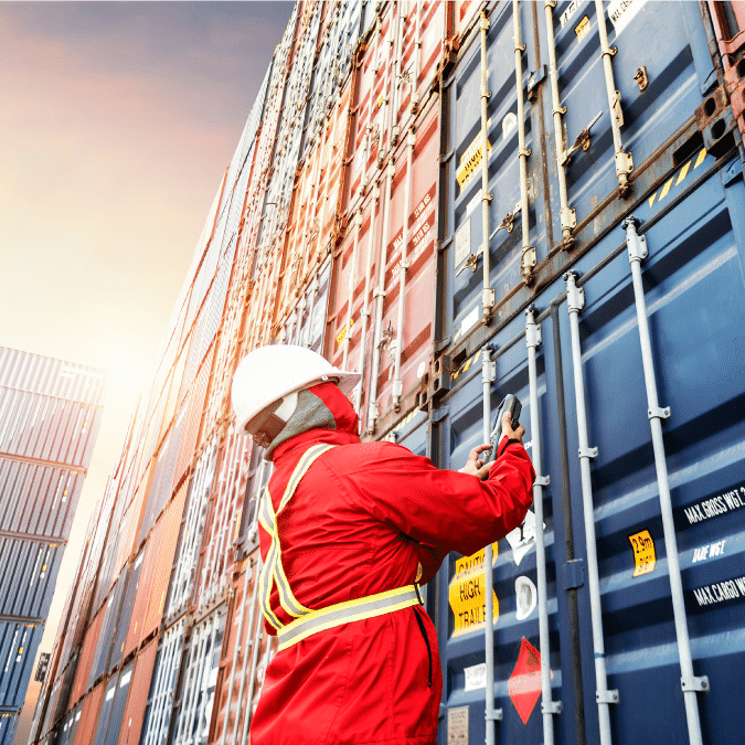 A worker wearing a red safety suit, helmet, and reflective vest inspects or secures stacks of shipping containers outdoors at a shipping yard, with sunlight illuminating the scene.