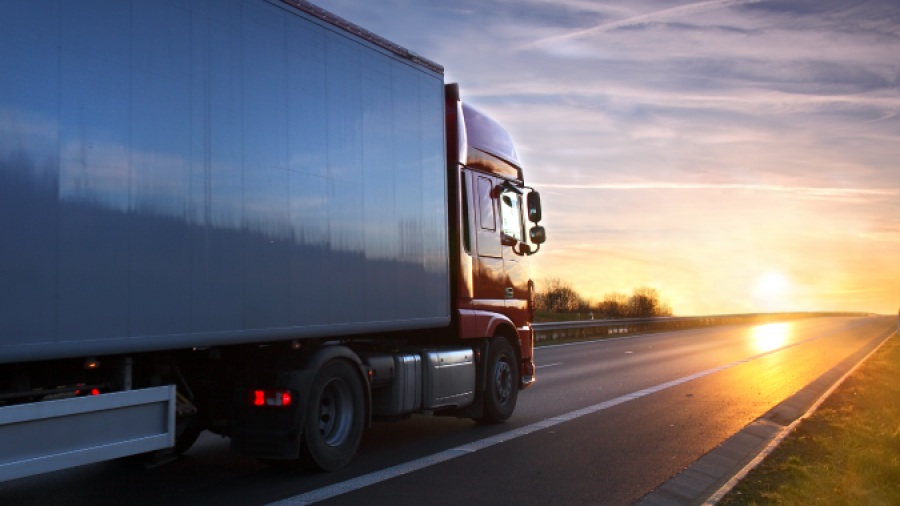 A large semi-truck drives down a highway at sunset, with the sun low on the horizon and a clear sky streaked with clouds and contrails.