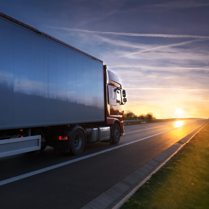 A large semi-truck drives down a highway at sunset, with the sun low on the horizon and a clear sky streaked with clouds and contrails.