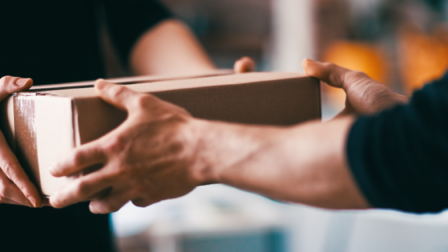 Two people’s hands exchanging a cardboard box indoors, suggesting a delivery or package handoff. The scene is focused on their arms and the box, with blurred background details.