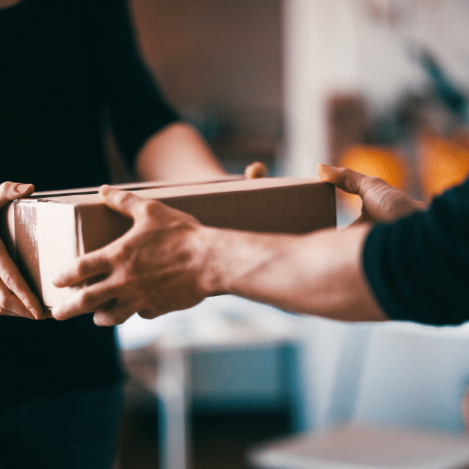 Two people’s hands exchanging a cardboard box indoors, suggesting a delivery or package handoff. The scene is focused on their arms and the box, with blurred background details.