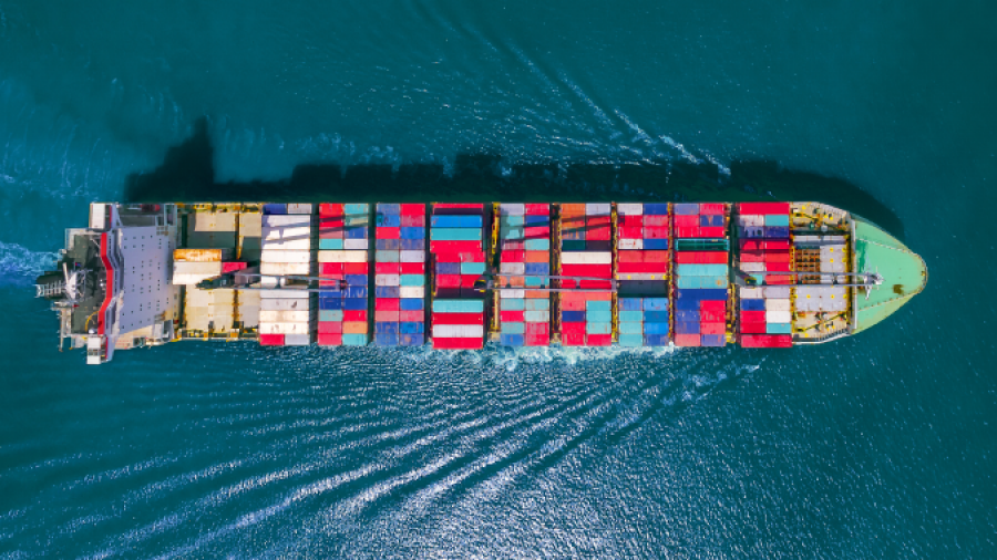 Aerial view of a large cargo ship carrying colorful shipping containers, sailing through deep blue ocean water and creating ripples behind it.