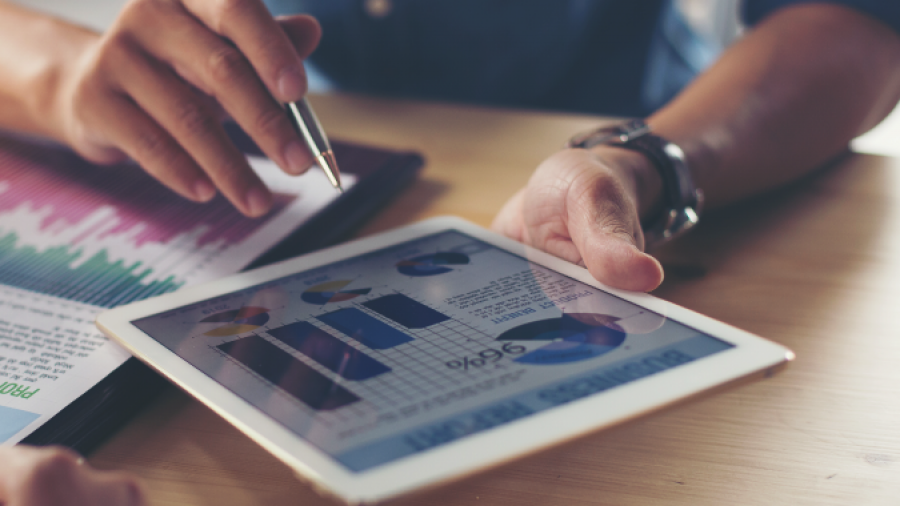 Two people sit at a table reviewing financial charts and graphs displayed on a tablet and printed paper, discussing data and analysis in a business setting.