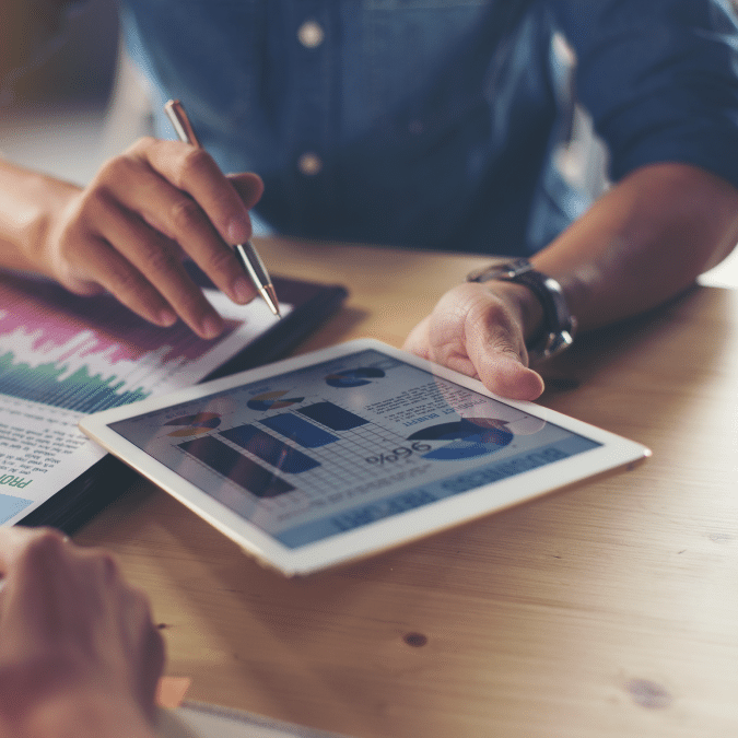 Two people sit at a table reviewing financial charts and graphs displayed on a tablet and printed paper, discussing data and analysis in a business setting.