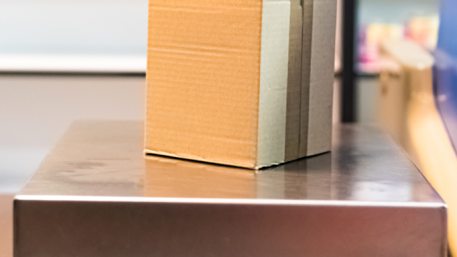A small cardboard box sits on a metal platform scale, likely in a shipping or postal setting, ready to be weighed.