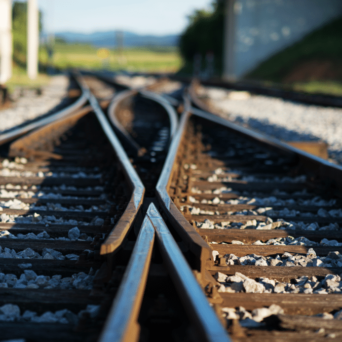 Close-up view of intersecting railway tracks with sunlight casting shadows, gravel surrounding the rails, and green scenery blurred in the background.