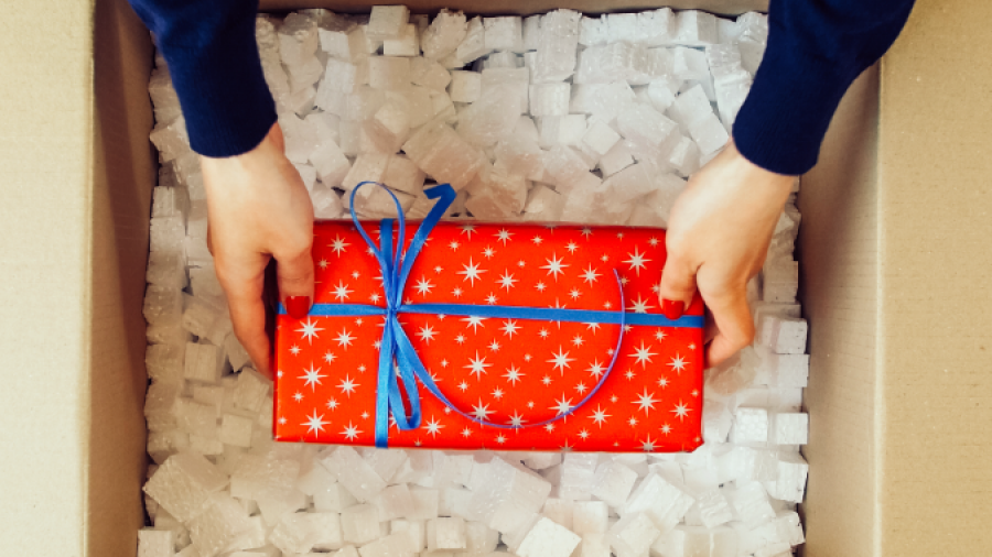 A person places a red gift wrapped with blue ribbon and white stars into a cardboard box filled with white packing peanuts.