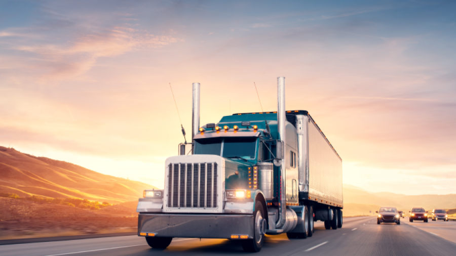 A large semi-truck drives down a highway at sunset, with other vehicles in the background and hills visible to the side under a colorful sky.