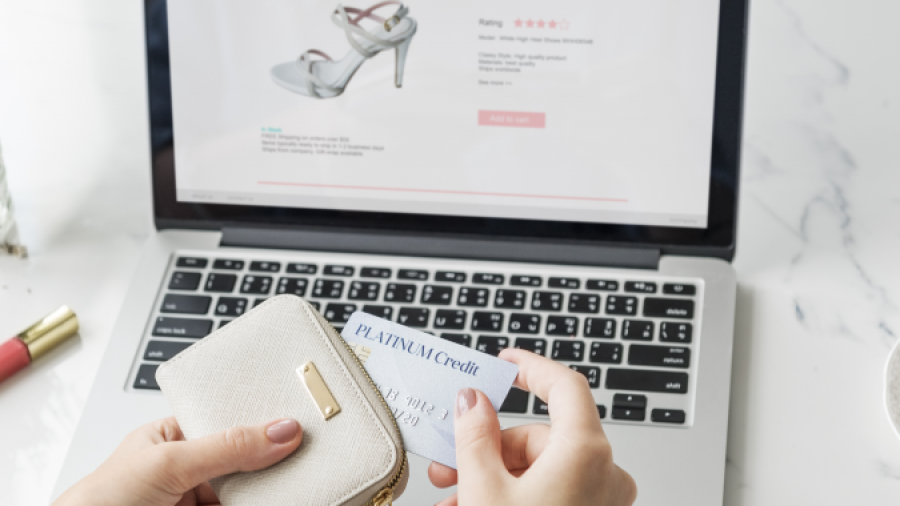 A person holds a beige wallet and a credit card while shopping for shoes online on a laptop. The laptop screen displays a sandal with reviews and a purchase option.