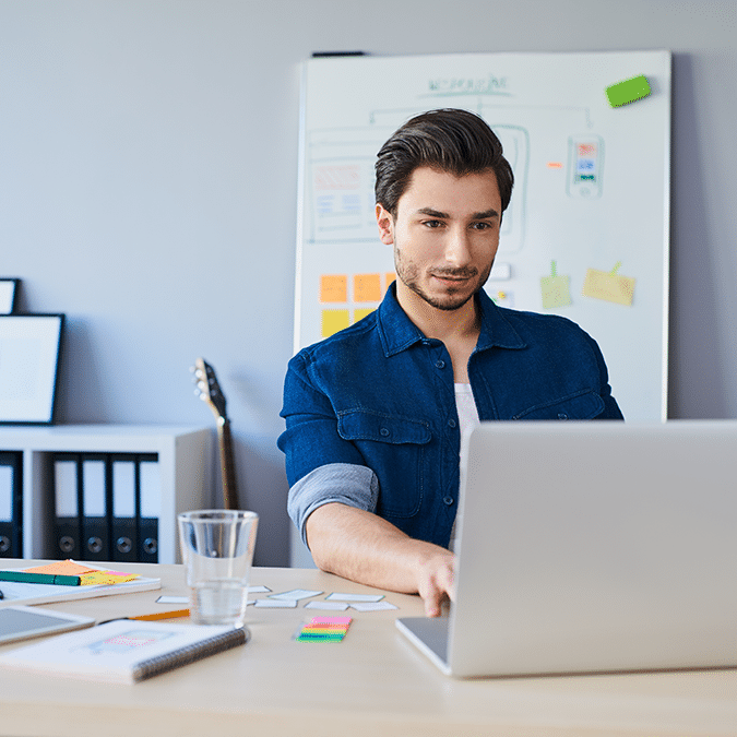 A man with dark hair and a beard works on a laptop at a desk in a modern office, with documents, a glass of water, and stationery nearby. A whiteboard with sketches is visible in the background.