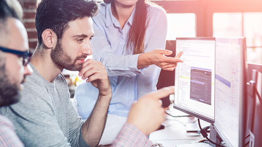 Three people collaborate in a bright, modern office. One woman stands, pointing at a monitor displaying Microsoft Power Apps, while two men sit and attentively discuss the app’s features and code on the screen.