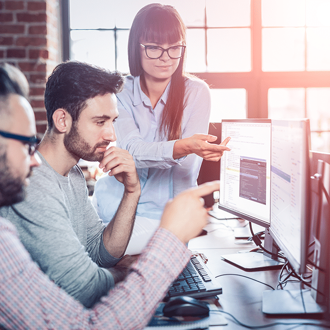 Three people collaborate in a bright, modern office. One woman stands, pointing at a monitor displaying Microsoft Power Apps, while two men sit and attentively discuss the app’s features and code on the screen.