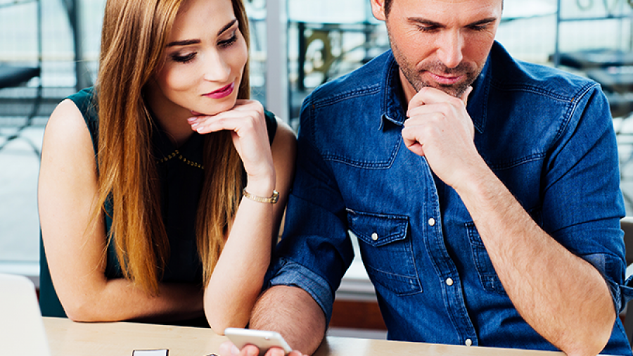 A man and woman sit at a desk, thoughtfully looking at cards with colorful labels. The man holds a phone, and a laptop is open beside them. They appear to be discussing or planning something together.