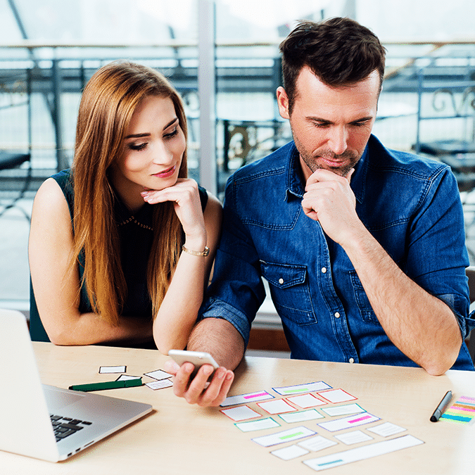 A man and woman sit at a desk, thoughtfully looking at cards with colorful labels. The man holds a phone, and a laptop is open beside them. They appear to be discussing or planning something together.