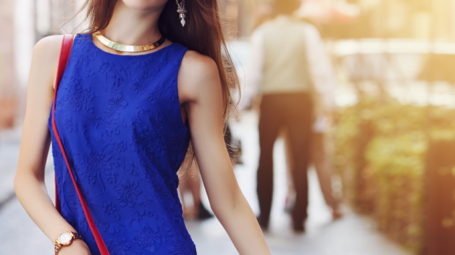 A stylish woman in a red hat and blue sleeveless top walks down a sunlit city street, carrying a red purse and wearing white pants, with blurred pedestrians and buildings in the background.