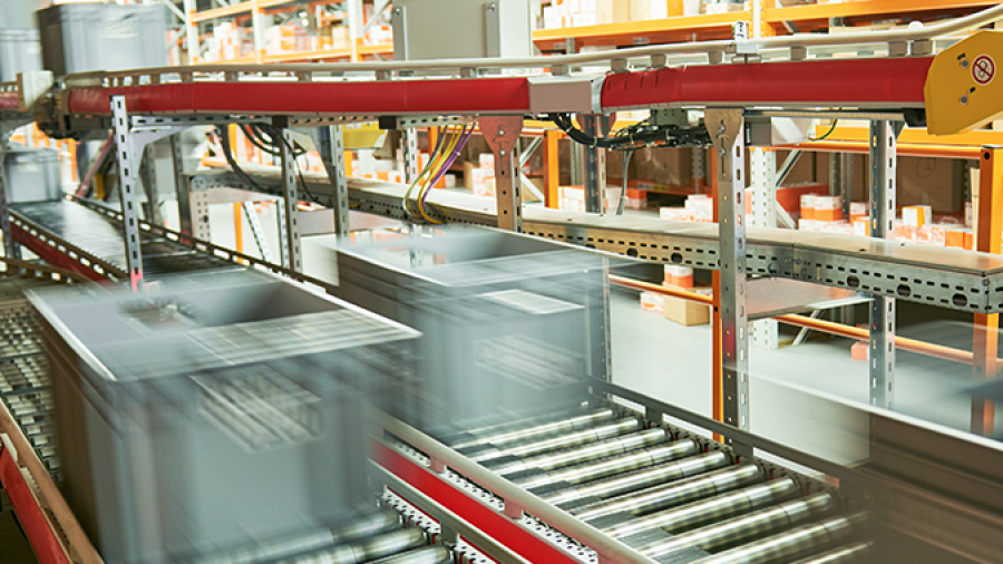 A warehouse conveyor system, designed with automation system engineering, transports large boxes rapidly across metal rollers, with shelves of orange and white packages in the background. The movement of the boxes appears blurred.
