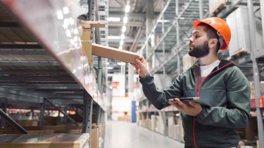 A man wearing an orange hard hat and green jacket holds a tablet while placing a box on a high shelf in a large warehouse filled with shelves and storage boxes.