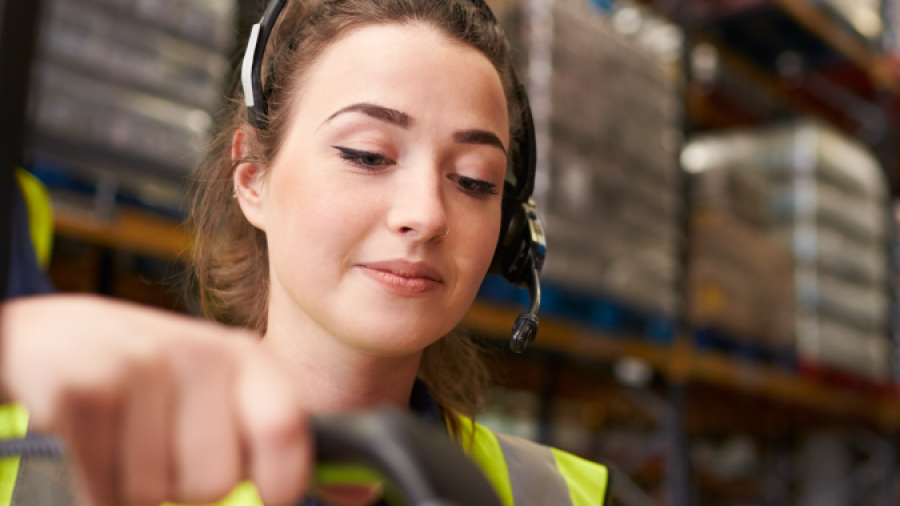 A warehouse worker wearing a headset and high-visibility vest scans a package with a barcode scanner in a storage facility with shelves full of boxes in the background.